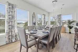 A dining area with a large table set for six, surrounded by chairs, with large windows and a view of the outdoors.
