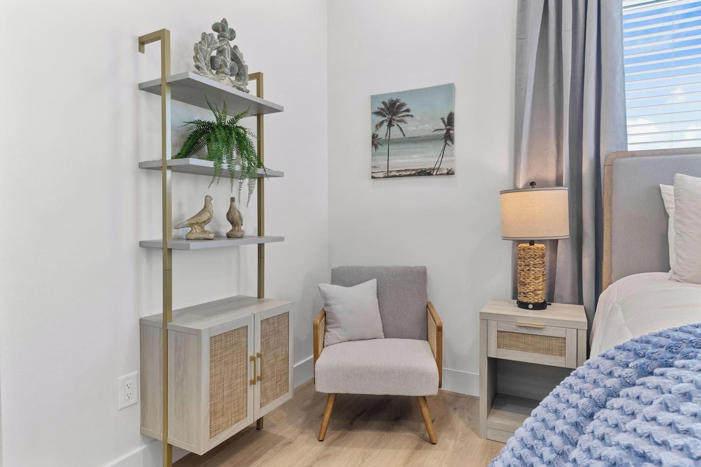 A bedroom with a bed, a chair, a nightstand, a lamp, a shelf with decorative items, and a painting of a beach scene.