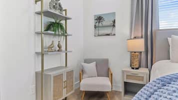 A bedroom with a bed, a chair, a nightstand, a lamp, a shelf with decorative items, and a painting of a beach scene.