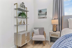 Un dormitorio con una cama, una silla, una mesita de noche, una lámpara, un estante con objetos decorativos y un cuadro de una playa.