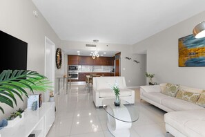 A modern living room with a glass coffee table, white sofas, and a dining area with a kitchen in the background. - ICON BRICKELL IMPRESSIVE 5★ OCEAN/POOL VIEWS @45th (Miami)
