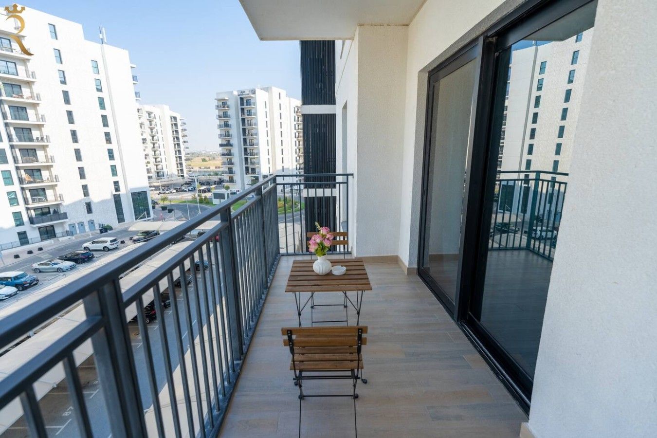 A balcony with a table and chairs, a vase with flowers, and a view of a cityscape with buildings and parked cars.