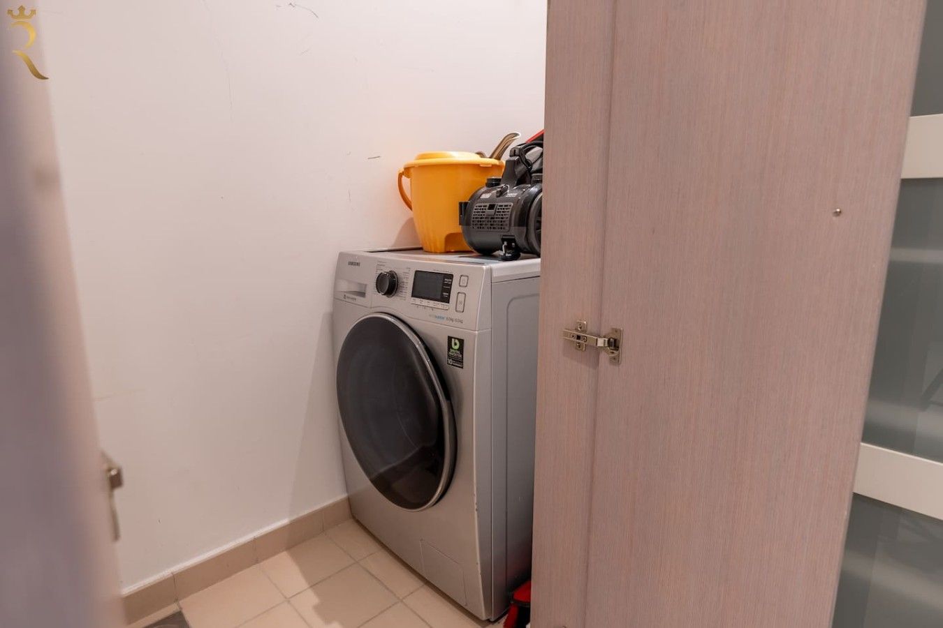 A compact laundry area with a washing machine, a yellow bucket, and a red container.