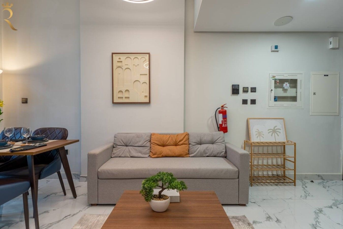 A modern living room with a sofa, a coffee table with a plant, a dining area with chairs and a table, and a wall-mounted fire extinguisher.