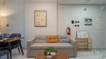 A modern living room with a sofa, a coffee table with a plant, a dining area with chairs and a table, and a wall-mounted fire extinguisher.