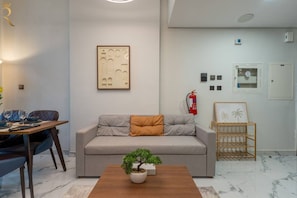 A modern living room with a sofa, a coffee table with a plant, a dining area with chairs and a table, and a wall-mounted fire extinguisher. - 1BR Sea View | Private Balcony | Maryah Vista (Abu Dhabi)