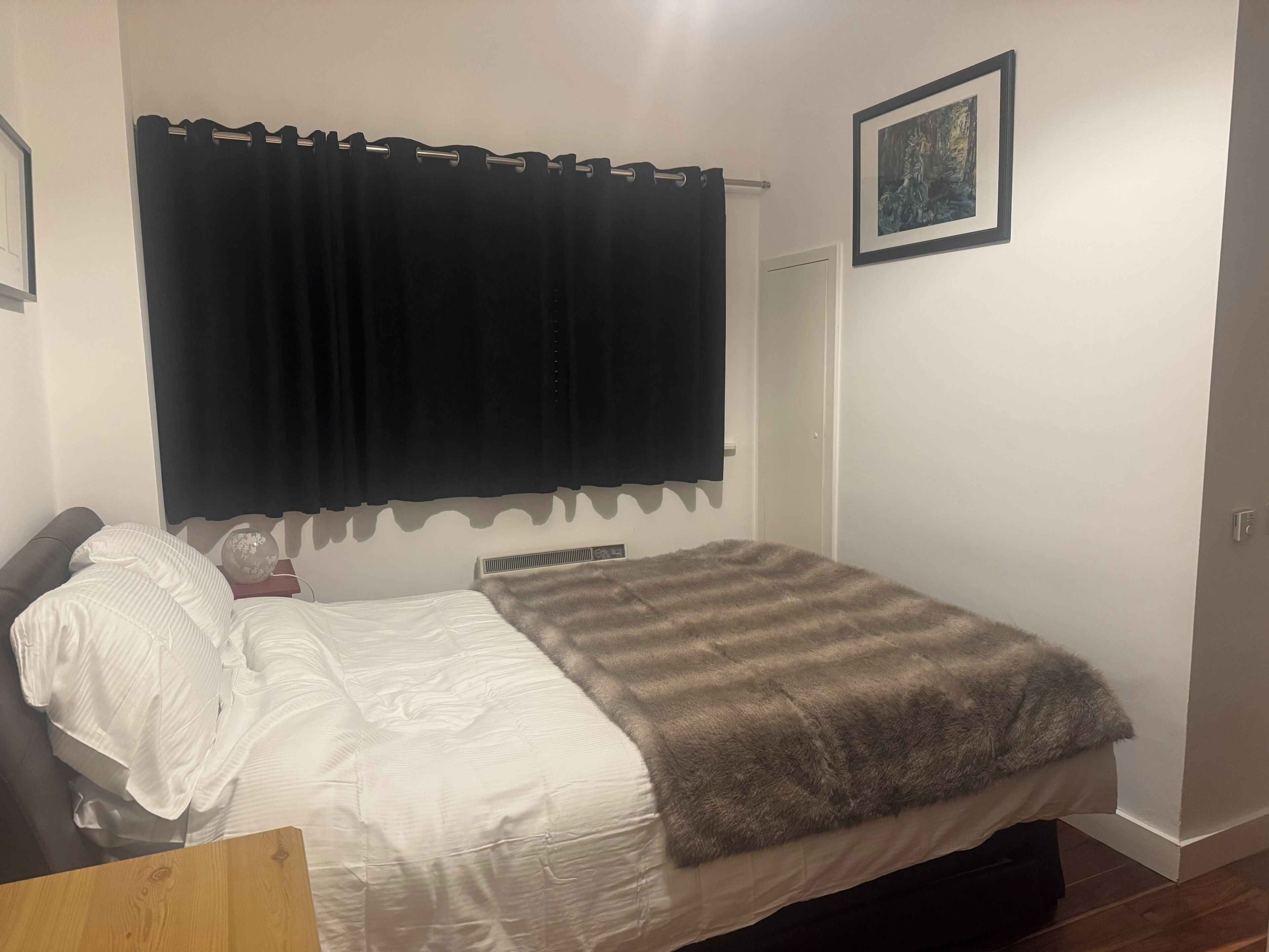 A bedroom with a bed, a bedside table, a heater, a curtain, a door, and a framed picture on the wall.