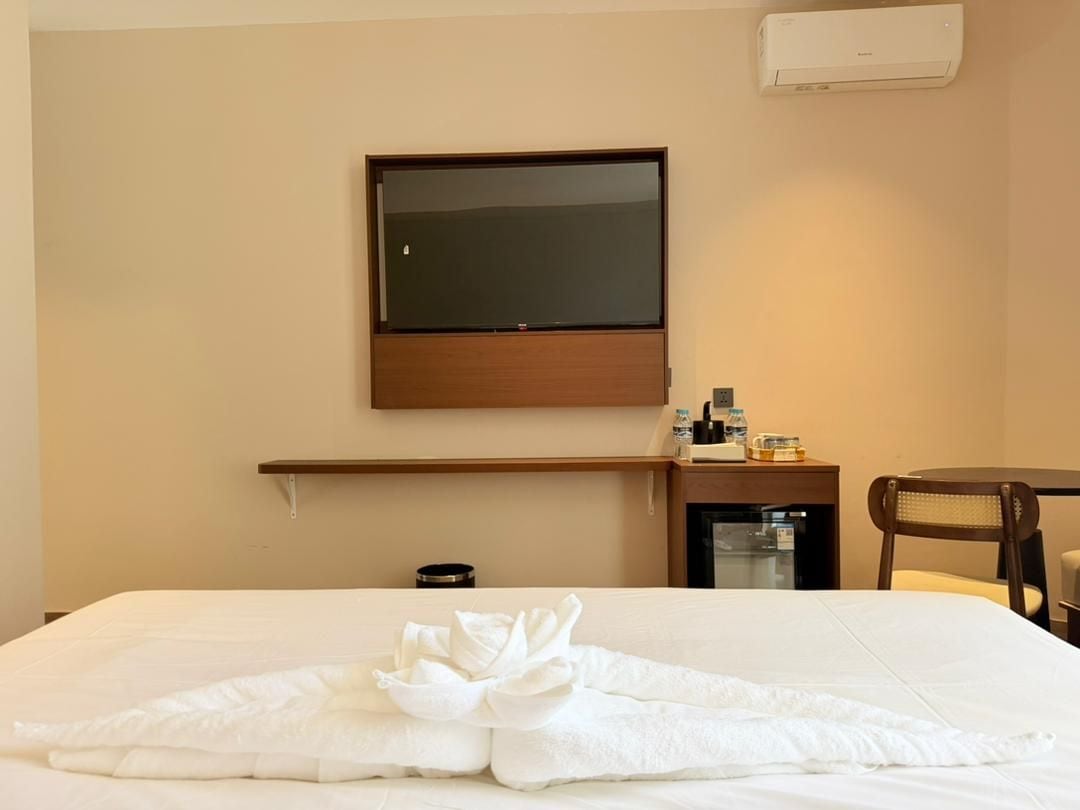 A hotel room with a bed, a flat-screen TV mounted on the wall, a small table with toiletries, and a chair.