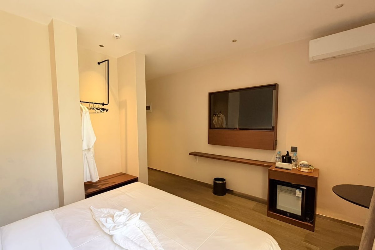 A hotel room with a bed, a mirror, a small table with a TV, and a towel rack.