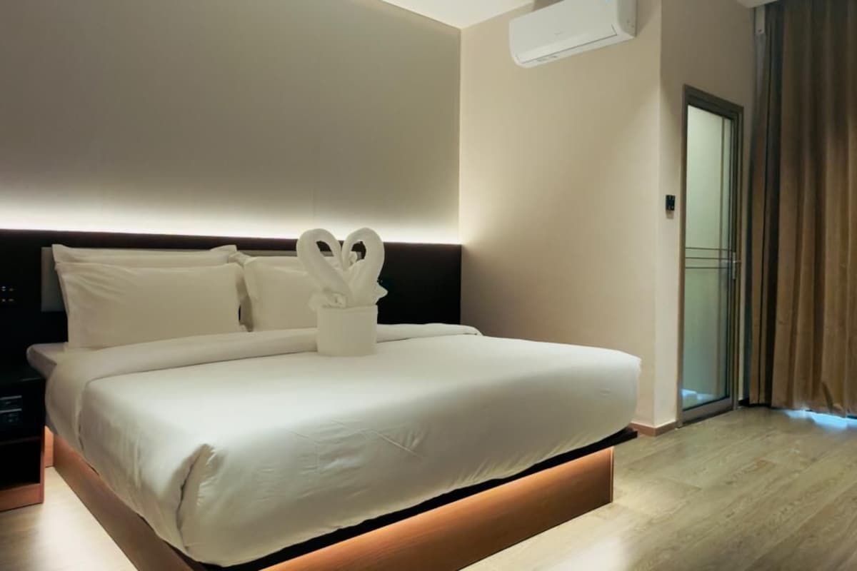 A modern hotel room with a large bed, a bedside table, an air conditioning unit, and a view of the outside through a glass door.