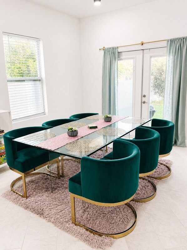 A dining area with a glass-top table, teal upholstered chairs, and a pink rug.