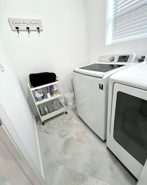 A laundry room with a wall-mounted sign, a white laundry cart with cleaning supplies, and a front-loading washer and dryer. - The Velvet Retreat | Glam Escape w/ Beauty Lounge (Tampa)