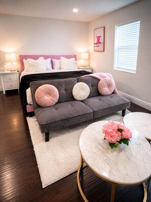 A bedroom with a bed, a grey sofa, a white rug, a round marble table, and a window with blinds. - The Velvet Retreat | Glam Escape w/ Beauty Lounge (Tampa)