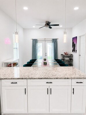 A modern kitchen with white cabinets, a granite countertop, a ceiling fan, and a dining area with a glass table and teal chairs. - The Velvet Retreat | Glam Escape w/ Beauty Lounge (Tampa)