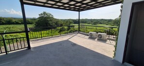 A covered balcony with a view of a green landscape, two air conditioning units, and a metal railing.