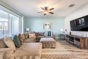 A living room with a sectional sofa, a ceiling fan, a flat-screen TV, and a large window. - Enchanting 6BR Vacation Home w/Pool @Championsgate (Davenport)