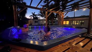 A hot tub with two people, surrounded by a wooden pergola and palm trees.