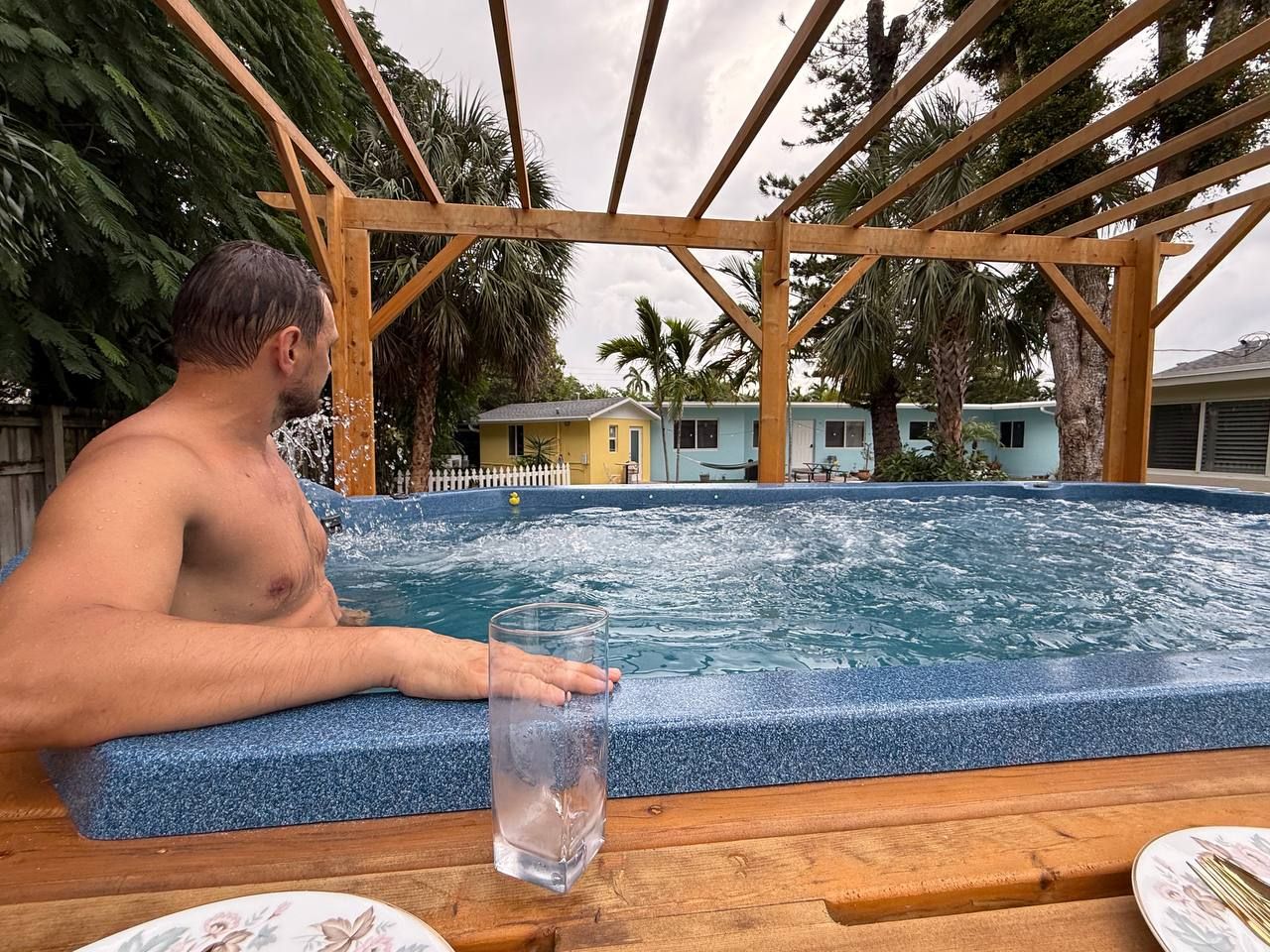 A person sitting by a hot tub with a clear glass of water, surrounded by a wooden pergola and palm trees.