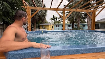 A person sitting by a hot tub with a clear glass of water, surrounded by a wooden pergola and palm trees.