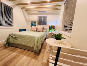 A bedroom with a bed, a bedside table with a lamp, a potted plant, and a window with blinds.