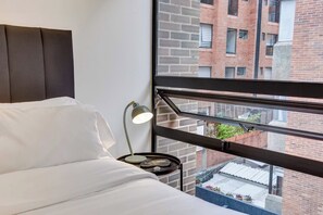 A hotel room with a bed, a nightstand with a lamp, and a window with a view of a brick building. - w* | Enchanting & Cozy Loft in the Heart of Bogotá (Bogotá)