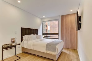 A bedroom with a bed, a bedside table with a lamp, a TV mounted on the wall, and a window with a view of a brick building. - w* | Enchanting & Cozy Loft in the Heart of Bogotá (Bogotá)