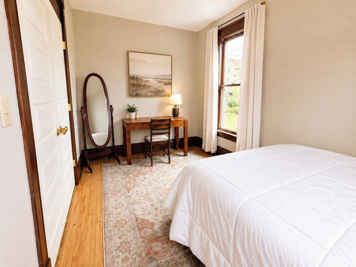 A bedroom with a bed, a desk with a chair, a mirror, a lamp, and a window with curtains.