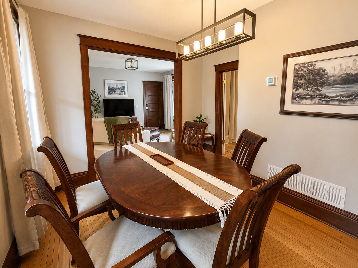 A dining room with a round wooden table, six chairs, a woven placemat, and a pendant light fixture.