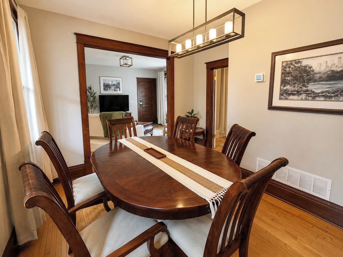 A dining area with a round wooden table, six chairs, a central rug, and a framed picture on the wall.