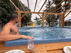Una persona sentada junto a un jacuzzi, con un vaso de agua cristalina, rodeada por una pérgola de madera y palmeras.