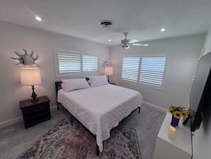 Un dormitorio con una cama, dos mesitas de noche con lámparas, un ventilador de techo y un televisor.