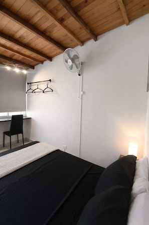 Un dormitorio con cama, escritorio, silla, perchero y ventilador de pared.