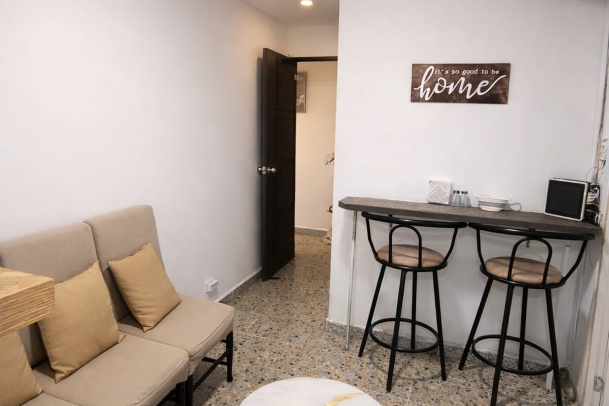 Un ambiente compatto con un divano, un tavolino rotondo, un angolo cottura con uno sgabello da bar e un cartello a parete.