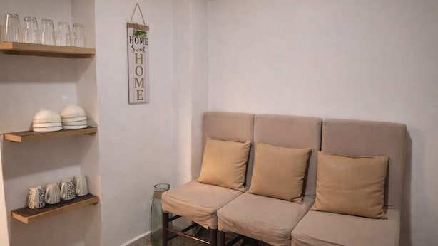 Ett rum med en neutral färgskala, en beige soffa, två hyllor i trä med glasföremål och dekorativa detaljer samt en väggmonterad skylt med texten ”Härliga hem”.