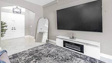 A modern living room with a large flat-screen TV mounted on the wall, a white entertainment unit below, and a patterned rug on the floor.