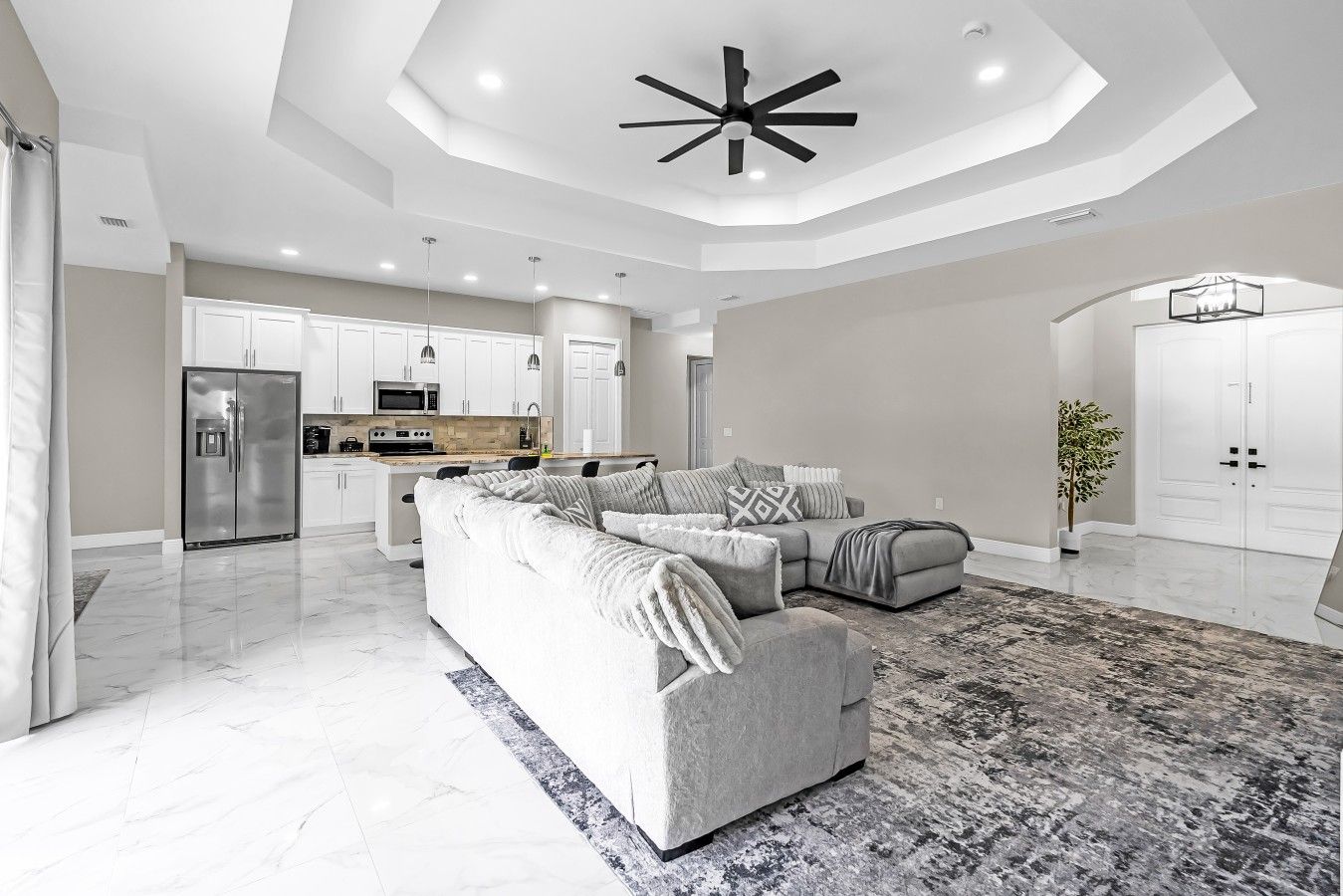 A modern living room with a sectional sofa, a dining area with a kitchen in the background, and a large ceiling fan.