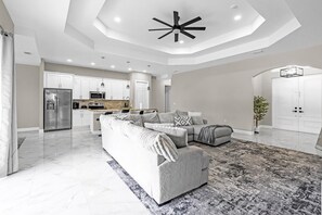 A modern living room with a sectional sofa, a dining area with a kitchen in the background, and a large ceiling fan. - Spacious 4BR Retreat, Lehigh Acres (Lehigh Acres)