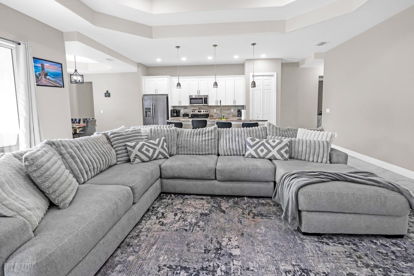 A modern living room with a sectional sofa, a kitchen in the background, and a large window.