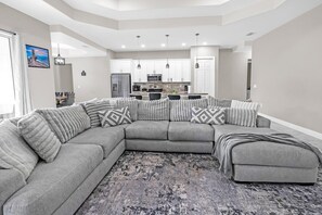 A modern living room with a sectional sofa, a kitchen in the background, and a large window. - Spacious 4BR Retreat, Lehigh Acres (Lehigh Acres)