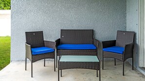 Ein Set aus Korbmöbeln mit blauen Kissen, darunter ein Sessel und ein kleiner Tisch, steht auf einer Beton-Terrasse vor einer strukturierten Wand.