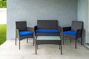 A set of wicker furniture with blue cushions, including a chair and a small table, positioned on a concrete patio against a textured wall. - Spacious 4BR Retreat, Lehigh Acres (Lehigh Acres)
