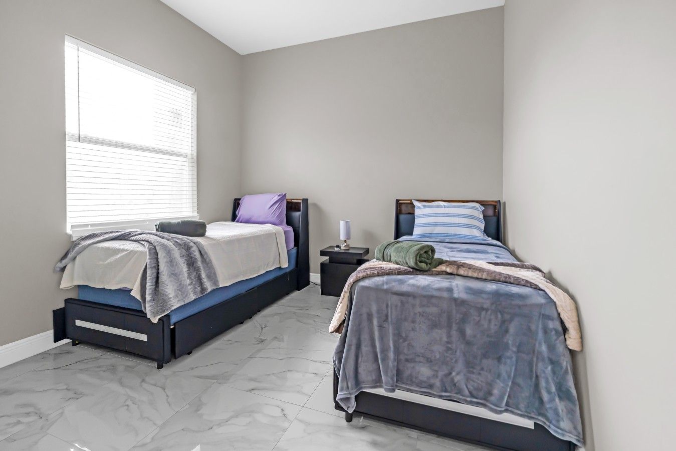 A bedroom with two beds, a nightstand, a lamp, and a window with blinds.