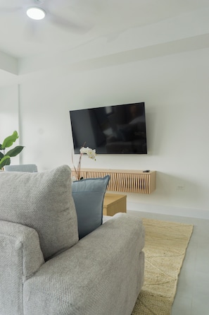 Una sala de estar moderna con un televisor de pantalla plana montado en la pared, un sofá gris, un cojín azul, una mesa consola de madera y una alfombra tejida.