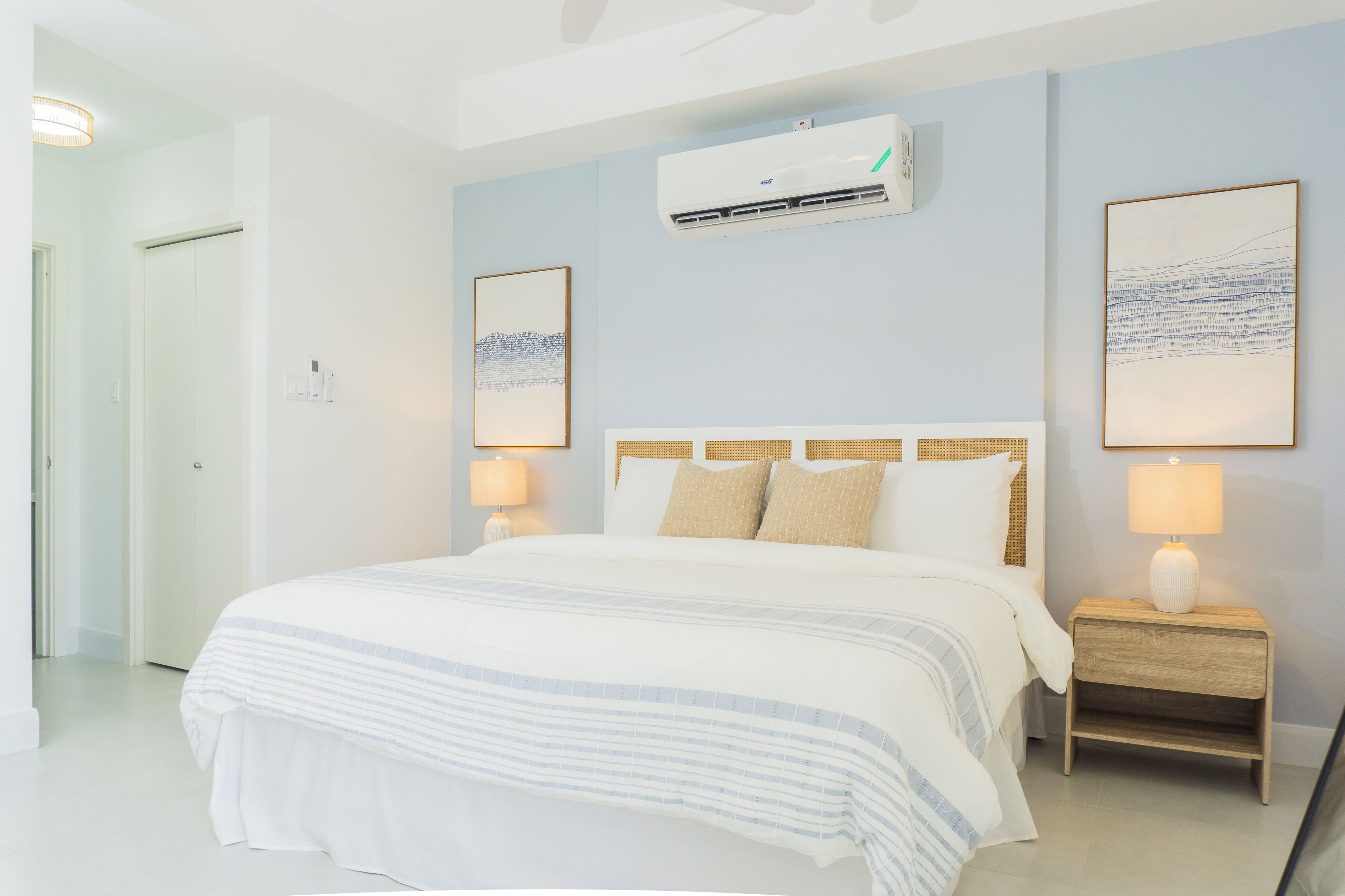 A bedroom with a bed, bedside lamps, an air conditioning unit, and two framed artworks on the wall.