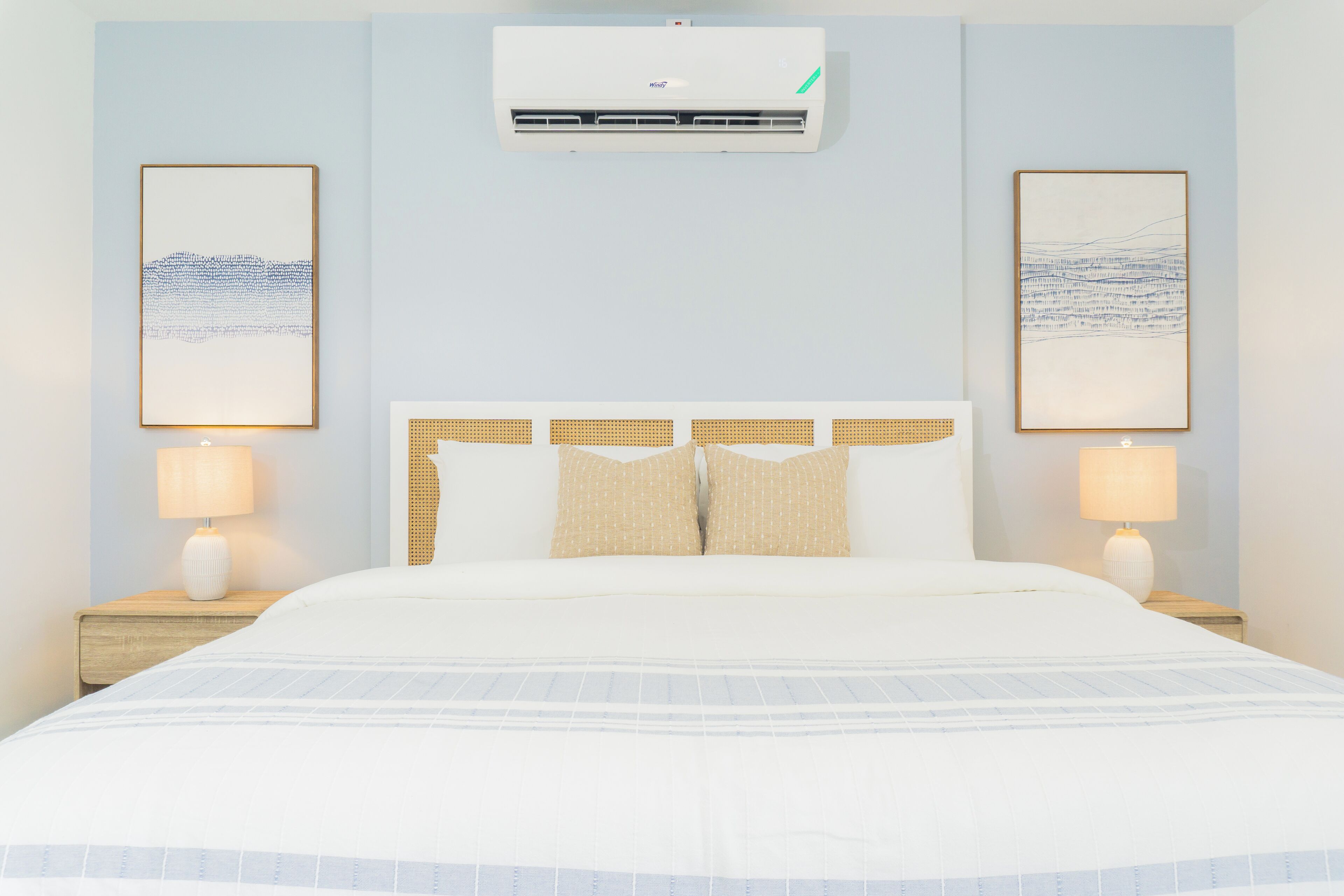 A hotel room with a bed, two bedside lamps, a wall-mounted air conditioner, and two framed artworks on the walls.