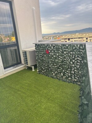 A rooftop terrace with artificial grass, a decorative wall with foliage, and a view of a cityscape.