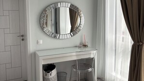 A room with a white desk, a round mirror, a clear chair, a door, and a window with curtains.