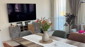 Moderni ruokasali, jossa on pitkä puinen pöytä, pehmustetut tuolit, maljakko täynnä kukkia ja seinälle asennettu litteänäyttöinen televisio.
