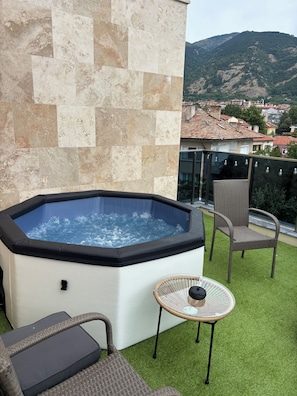 Banyera d'hidromassatge en un balcó amb gespa artificial, mobles de vímet i vista a un poble situat en un turó.