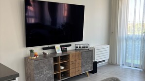 A modern living room with a flat-screen TV, a wooden cabinet with drawers, a dining table, and a large window with sheer curtains.